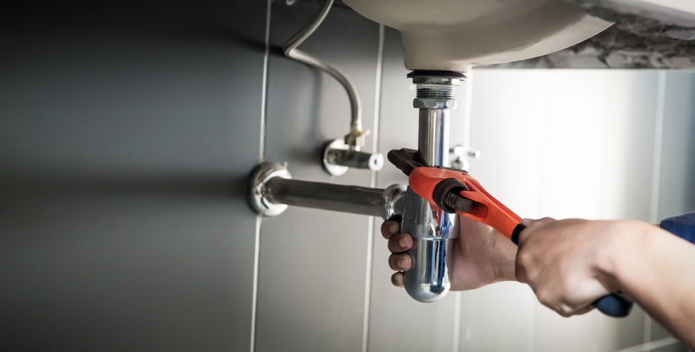 A plumber El Dorado County, CA, uses a red pipe wrench to tighten or loosen a shiny metal pipe under a bathroom sink. The background shows tiled walls and plumbing connections.