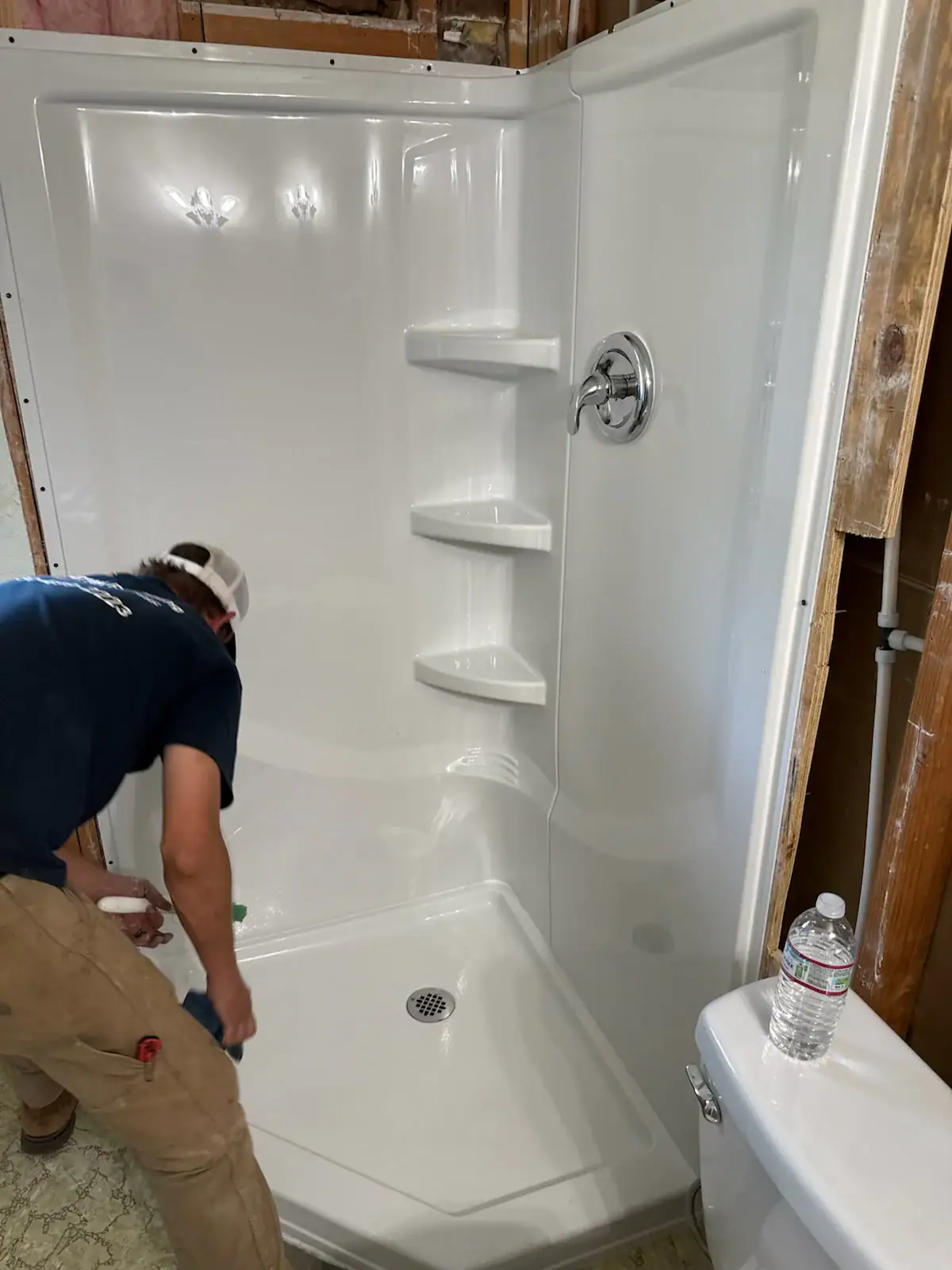 A person in a navy shirt and tan pants cleans a new white shower stall with shelves, next to exposed wooden wall studs and a white toilet with a water bottle on the tank.