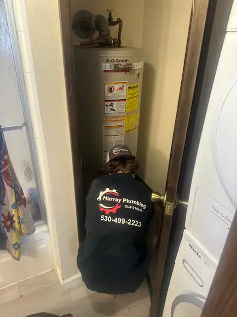 A plumber wearing a "Murray Plumbing" jacket is crouched in front of a water heater in a small closet, working on it. The plumber's back is to the camera. A washer and dryer are visible nearby.