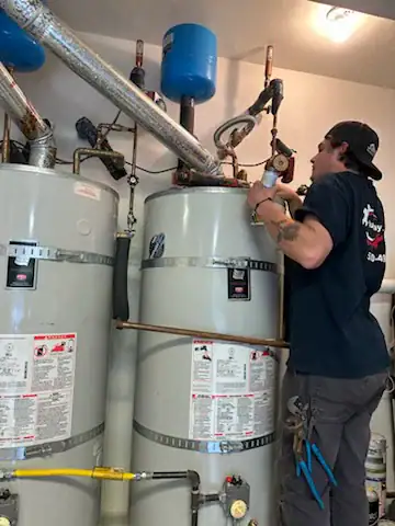 A technician wearing a black shirt works on a large water heater system, using tools to adjust pipes and valves connected to two tall cylindrical tanks in a utility room.