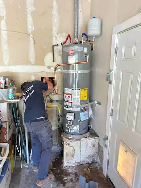 A person installs or repairs a water heater in a garage, working with tools near pipes and an unfinished wall. The water heater is mounted on a raised platform beside a door. Various items are stored around the workspace.