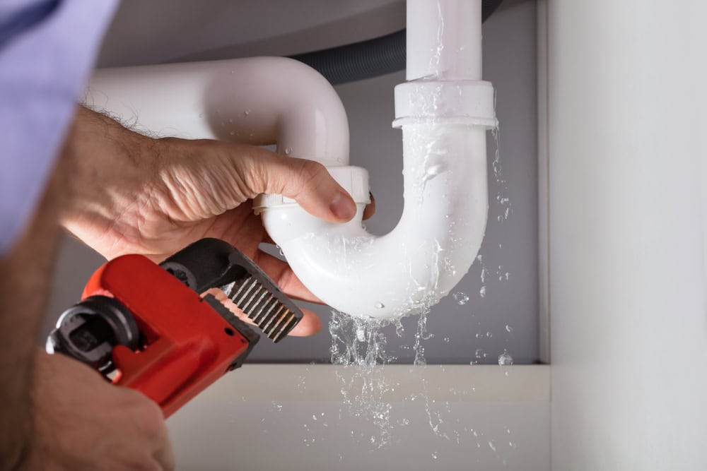 A plumber in El Dorado County uses a red pipe wrench to fix a leaking white pipe under a sink, with water spraying out from the joint.