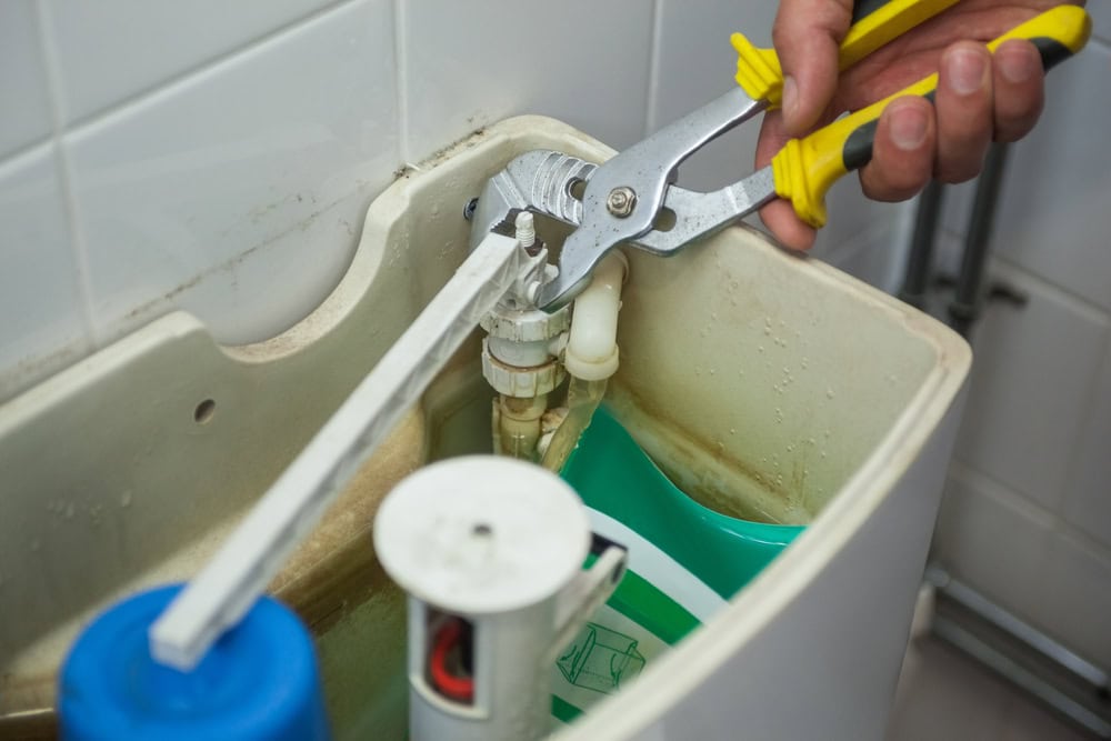 A plumber in El Dorado County, CA uses pliers to repair or adjust the mechanism inside a toilet tank with visible mineral stains and water lines.