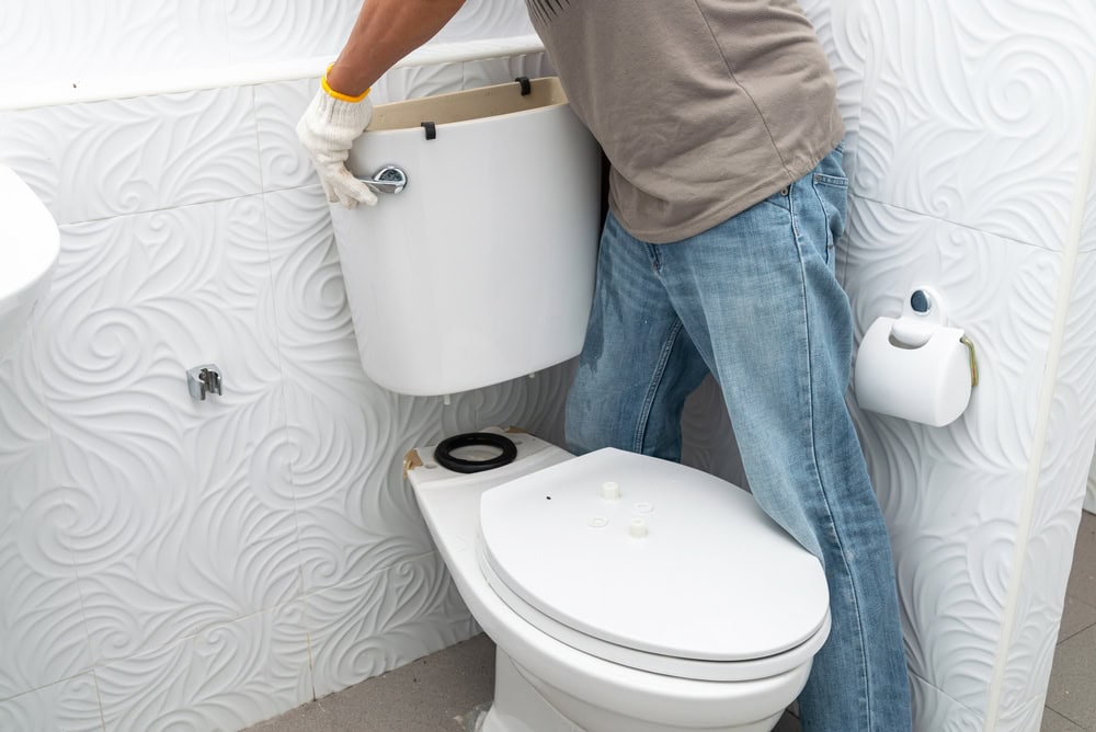 A plumber El Dorado County, CA, wearing gloves and jeans, is installing or repairing a toilet tank in a bathroom with white, textured swirl-patterned walls.