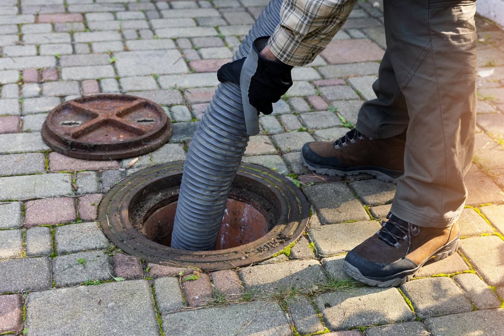 A plumber in El Dorado County, CA, wearing boots and gloves, uses a large hose to clean or empty an outdoor septic tank through an open manhole on a paved surface, with the manhole cover placed nearby.