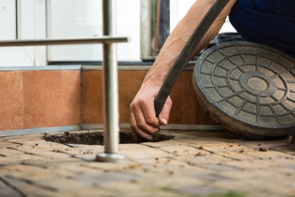 A plumber in El Dorado County, CA lifts a round metal manhole cover and inserts a hose into an outdoor drain on a tiled surface, possibly for cleaning or maintenance work.