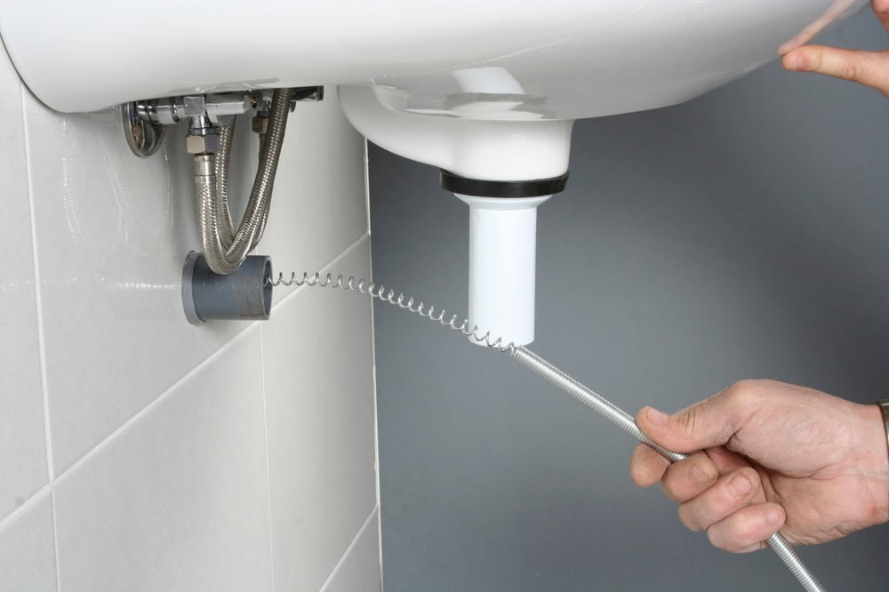 A plumber El Dorado County, CA is using a plumbing snake to unclog the pipe under a white sink attached to a tiled wall.