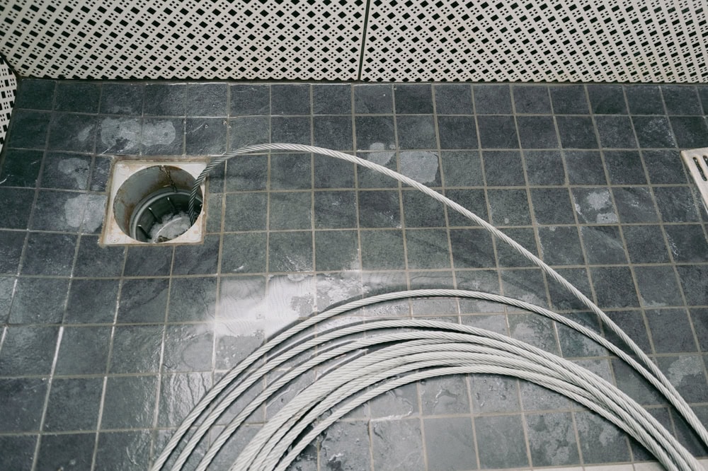 A coil of metal cable lies on a dark tiled floor next to an open drain surrounded by wet patches—just the kind of scene a plumber El Dorado County, CA expert might encounter during water heater replacement or repairs. White patterned tiles line the walls in the background.