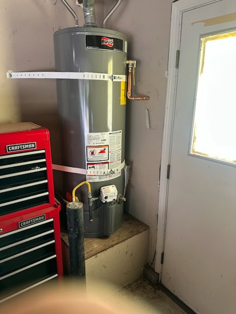 A gray Ruud water heater is installed on a raised platform in a CA garage next to a red Craftsman tool chest and near a door with a window. Secured with metal straps, it’s an example of quality plumber El Dorado County installation.