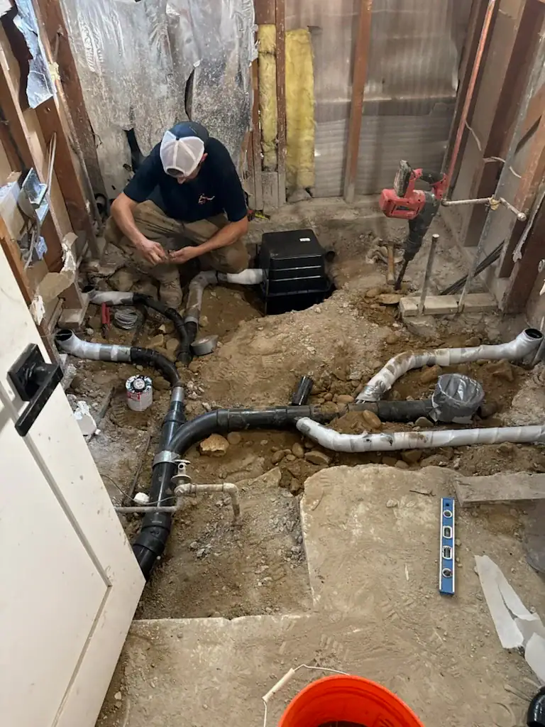 A plumber kneels in a partially demolished bathroom, working on exposed black PVC pipes and drains in the floor. Tools, a level, and construction materials are scattered around the unfinished space in El Dorado County, CA