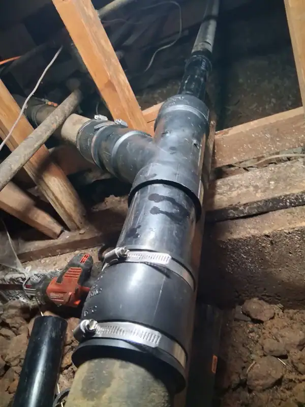 A close-up view of black PVC pipes with metal clamps and fittings installed in a crawl space with exposed wooden beams and dirt floor. A power tool is visible to the left in El Dorado County, CA