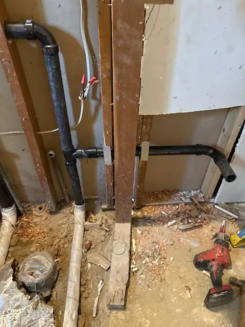 A partially demolished wall reveals exposed black plumbing pipes, electrical wiring, wood framing, construction debris, and a red cordless power drill lying on the dusty floor in El Dorado County, CA