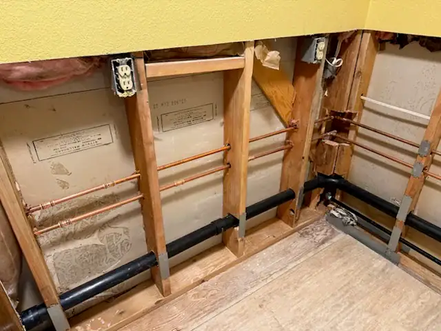 Exposed wall studs with visible plumbing pipes, electrical outlets, and insulation in a partially renovated room with a plywood floor and yellow-painted upper wall in El Dorado County, CA