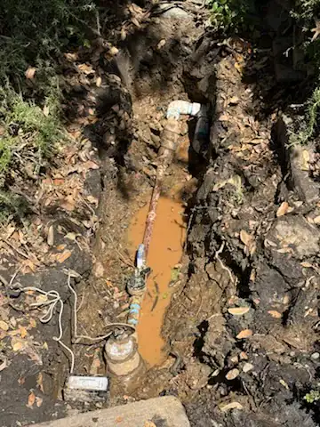 A muddy trench with exposed water pipes and valves, partially filled with murky brown water, surrounded by soil, roots, and scattered leaves in El Dorado County, CA