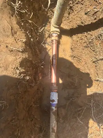 A section of copper pipe with fittings is installed in a trench with exposed soil and roots, connecting to two other pipes. Shadows partially cover the area in El Dorado County, CA