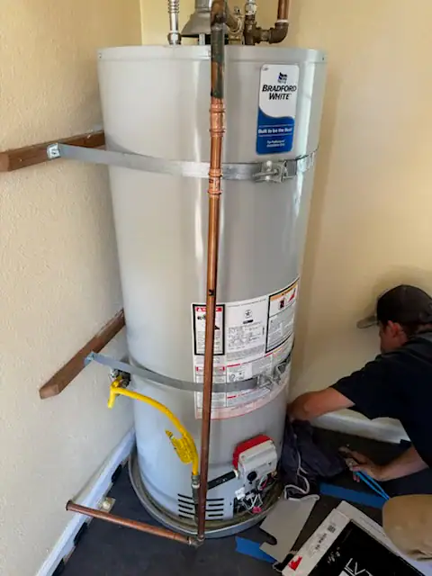 A person works on installing or repairing a Bradford White water heater in the corner of a room. Various pipes, wires, and tools are visible around the unit, and the water heater is secured to the wall with metal straps.