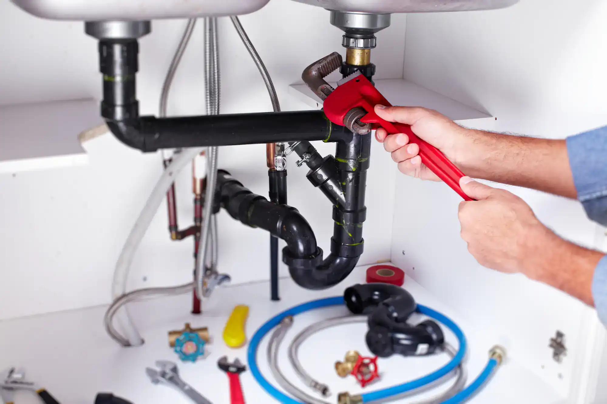 A person uses a red pipe wrench to tighten a pipe under a sink; various plumbing tools and supplies are spread out on the cabinet floor in El Dorado County, CA
