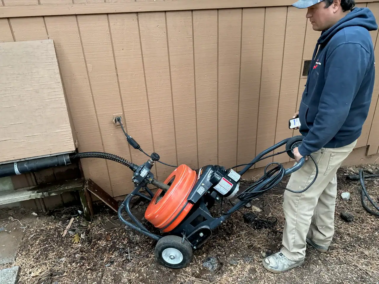 A person operates a sewer drain cleaning machine with an orange reel near an exterior wooden wall, feeding the cable into a pipe opening in El Dorado County, CA