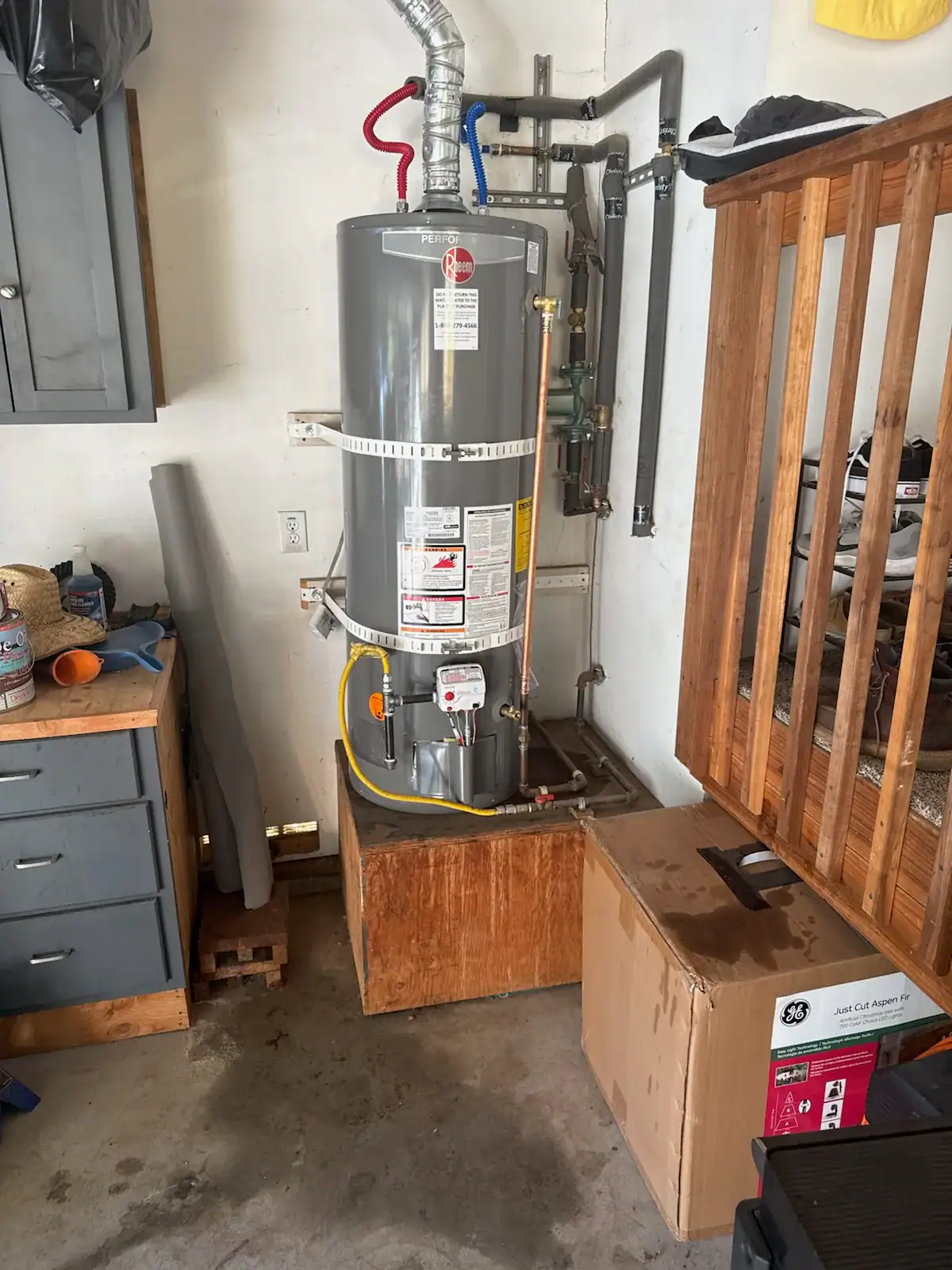 A gray water heater is installed in a corner of a garage, with pipes connected to the top. Nearby are a cardboard box, a wooden platform, and a workbench with tools and items on it in El Dorado County, CA