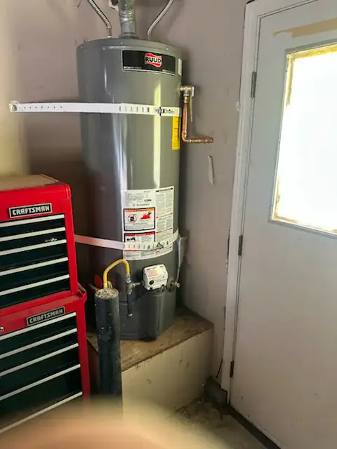 A gray Ruud water heater is installed on a raised platform in a garage next to a red Craftsman tool chest and a door with a window. The water heater is secured with white straps in El Dorado County, CA